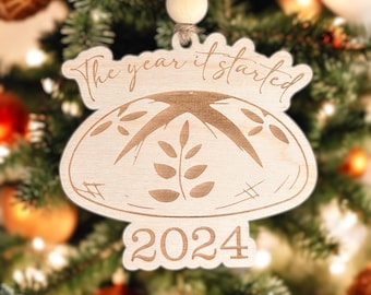 May include: A wooden Christmas ornament shaped like a sourdough bread loaf with the text "The year it started 2024" on it.