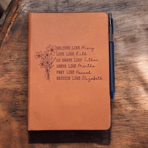May include: A tan-colored notebook with the words "BELIEVE LIKE Mary, LOVE LIKE Ruth, BE BRAVE LIKE Esther, SERVE LIKE Martha, PRAY LIKE Hannah, REJOICE LIKE Elizabeth" and a floral illustration. A blue pen rests along the right side.