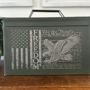May include: A green metal ammo can with a distressed American flag, an eagle, and the words "We the People" and "FREEDOM" printed on the side.