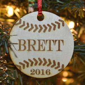 Op de afbeelding: Ronde houten kerstversiering met een honkbalthema. De lichtbruine ornament heeft gegraveerde honkbalsteken, de naam "BRETT" en het jaar "2016". Een rood lint is bevestigd om op te hangen.