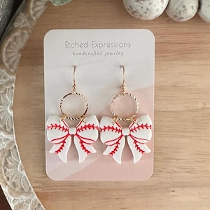 May include: White and red baseball-themed bow earrings with gold hoops. The earrings are on a white card with the text "Etched Expressions handcrafted jewelry".