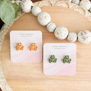 May include: Two pairs of frog earrings on pink cards. The cards have the text "Etched Expressions handcrafted jewelry". One pair of earrings is orange and the other pair is green.