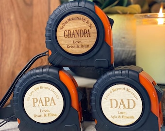 Könnte beinhalten: Drei schwarze und orangefarbene Maßbänder mit Holzeinsätzen. Die Einsätze haben personalisierte Botschaften für "Opa", "Papa" und "Vater" mit den Namen der Schenker.