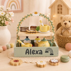 Può includere: Un supporto per set da tè giocattolo in legno a due livelli, color verde salvia, con un arco floreale. La base è personalizzata con il nome Alexa e un disegno di un arcobaleno. Include tazze, una teiera e pasticcini in legno, con biscotti.