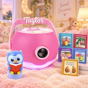 May include: A pink spherical children's storybook projector with a handle labeled Taylor and a Luna logo. It features a front lens, a glowing base, and a blue owl figurine holding a red book. Five small storybooks are stacked nearby.