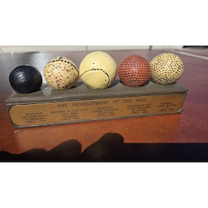 Könnte beinhalten: Ein Ausstellungsset mit dem Titel THE DEVELOPMENT OF THE BALL, das fünf historische Golfbälle auf einem rechteckigen Sockel zeigt. Die Bälle umfassen einen schwarzen Holzball, einen gesprenkelten Lederball, einen glatten cremefarbenen Ball, einen roten strukturierten Ball und einen Ball mit Vertiefungen.