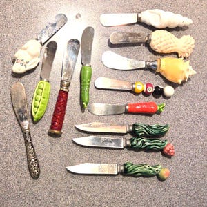 May include: A collection of twelve decorative spreaders and knives featuring varied handles including a white rabbit, a green pea pod, a red textured cylinder, a green vegetable, a white shell, a pineapple, a seashell, billiard balls, a carrot, and three green leafy designs with small fruit accents.