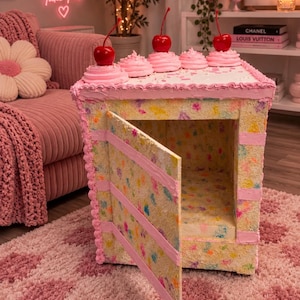 May include: A square cabinet designed to resemble a slice of cake with a confetti-patterned exterior, pink frosting trim, and three red cherry toppers on the top surface. The cabinet features a single hinged door that opens to reveal an interior.