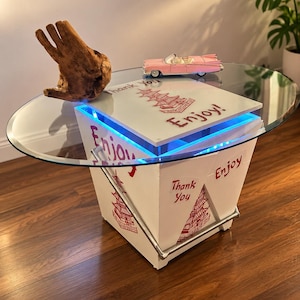 以下が含まれることがあります： テイクアウト容器の形をした白いコーヒーテーブル。赤いパゴダのグラフィックと「Thank You」「Enjoy」の文字が描かれています。ガラスの天板、金属製のハンドル、内部の青いLED照明が特徴です。木製のハンドスカルプチャーが上に置かれています。