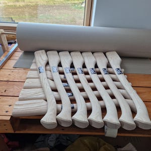 May include: A collection of seven unfinished light-colored wooden axe handles arranged in a row. Each handle features a curved, ergonomic shape with a visible wood grain pattern and a small rectangular black and white label attached to the surface.