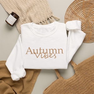 Puede incluir: Una sudadera blanca de cuello redondo con el texto Autumn Vibes en una fuente serif y cursiva de color tostado. La prenda se muestra junto a una manta texturizada beige, una pequeña vela en frasco marrón y un mantel individual redondo de mimbre.