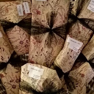 May include: Several rectangular packages wrapped in cream-coloured paper with a botanical print of brown and red wildflowers. Each package is tied with a black tulle ribbon and features a white label reading Mystery Doll and Not suitable for children.