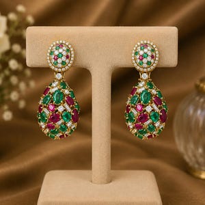 May include: A pair of gold drop earrings featuring a circular top and a larger pear-shaped bottom, both encrusted with a cluster of oval-cut green emeralds, oval-cut red rubies, and small round and baguette-cut white diamonds in a dense setting.
