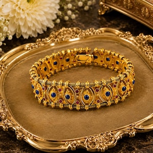 May include: A gold-toned metal bracelet featuring a repeating pattern of oval-shaped links. The bracelet is decorated with small, round, deep blue stones and smaller, round, vibrant red stones set within a textured, filigree-style metal frame.