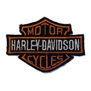 May include: A black and orange embroidered patch featuring the Harley-Davidson Motor Cycles logo. The design includes the text MOTOR at the top, HARLEY-DAVIDSON in white across the center bar, and CYCLES at the bottom, all in a shield shape.