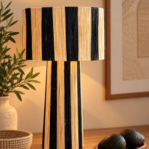 以下が含まれることがあります： 黒とナチュラルベージュの繊維を交互に縦縞模様で巻き付けた、円筒形のベースとドラム型のシェードを持つテーブルランプ。ランプは温かみのある光を放ち、巻き付けられた素材の質感を照らし出しています。