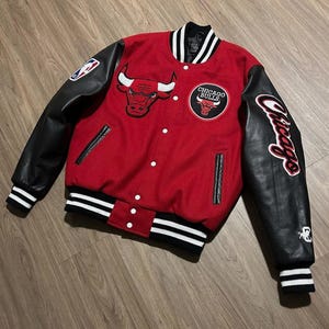Puede incluir: Una chaqueta universitaria roja con mangas de cuero negro, que presenta el logotipo de los Chicago Bulls en el pecho y la manga, botones a presión blancos y ribetes acanalados a rayas blancas y negras en el cuello, puños y dobladillo, con un parche NBA.