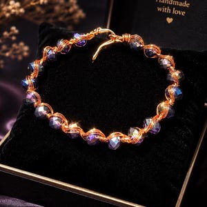 May include: A circular bracelet crafted from copper-coloured wire wrapped around faceted, iridescent purple beads. The bracelet features a simple hook-and-eye closure mechanism and is displayed on a black velvet cushion inside a box with the text Handmade with love.
