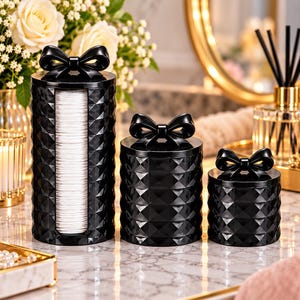 May include: A set of three black cylindrical containers with a diamond-textured surface and a bow-shaped handle on each lid. The tallest container features a vertical rectangular cutout displaying a stack of white circular cotton pads inside.