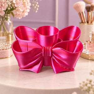 May include: A bright fuchsia desk organiser shaped like a large bow with a central cylindrical compartment and four surrounding curved sections. The surface has a smooth, metallic sheen and a distinct layered texture from the 3D printing process.