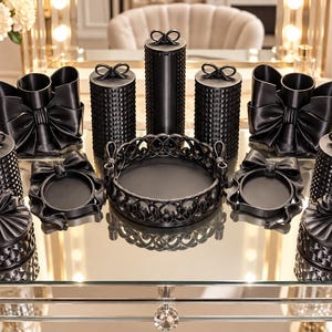 May include: A collection of black bathroom accessories including lidded canisters with textured surfaces, bow-shaped containers, circular trays with intricate lattice edges, and small round jars, all featuring decorative bow accents on the lids.