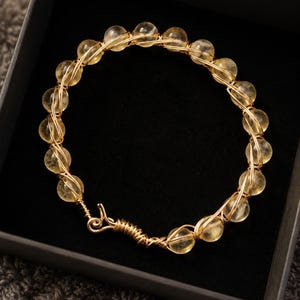 May include: A circular bracelet constructed from translucent yellow citrine beads wrapped in thin gold-toned wire. The bracelet features a hand-twisted wire hook-and-eye clasp mechanism, showcasing a continuous design of beads secured by wire.