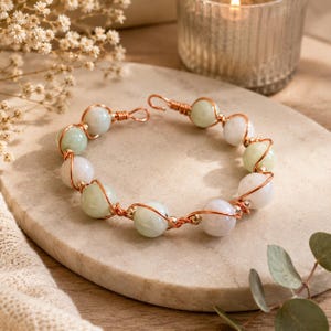 May include: A bracelet featuring round, pale green and white stone beads wrapped in copper wire. The wire forms a continuous spiral around each bead, connecting them in a circular chain. The bracelet includes a simple hook-and-eye clasp mechanism.
