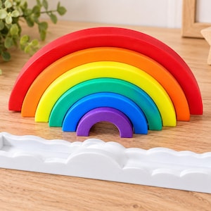 May include: A multi-colored stacking toy consisting of six nested semi-circular arches in red, orange, yellow, green, blue, and purple, paired with a white rectangular base featuring a scalloped cloud-like edge along the front and back.
