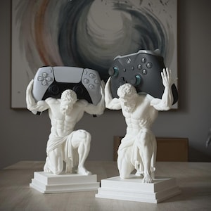 May include: Two white statues of kneeling figures with muscular builds, each mounted on a square base. The figures are positioned to hold gaming controllers on their shoulders, one featuring a white controller and the other a black controller.