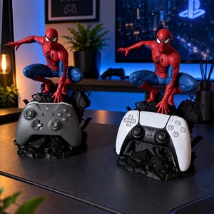 May include: Two Spider-Man action figures in a crouching pose, each mounted on a black, jagged, rock-like base. One figure is positioned above a grey wireless game controller, while the other is positioned above a white and black wireless game controller.