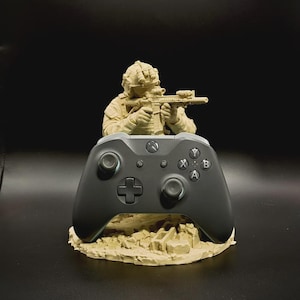 May include: A matte beige figurine of a soldier in tactical gear aiming a rifle, positioned behind a black wireless video game controller. The soldier stands on a textured, rocky base that supports the controller in an upright display position.