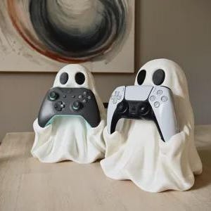 May include: Two white ghost-shaped controller stands made of a smooth material. The left ghost holds a black gaming controller, while the right ghost holds a white and black gaming controller. Both ghosts feature simple black oval eyes and draped fabric.