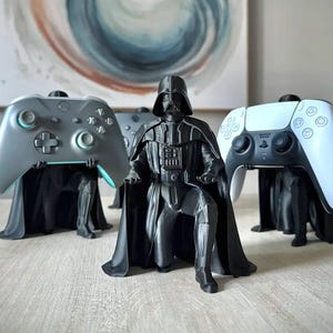 May include: Three black 3D-printed Darth Vader figures in a kneeling pose, each serving as a controller stand. Two figures hold gaming controllers, one grey and one white, while the central figure is empty, showcasing the detailed black armour.