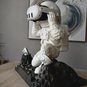 May include: A white astronaut figurine kneeling on a black cratered lunar surface base. The astronaut wears a white space suit and holds a white virtual reality headset over its helmet. Two white VR controllers are mounted into the black base.
