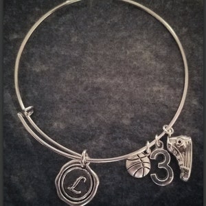 May include: Silver-toned bangle bracelet with multiple charms. Charms include a basketball, the number 3, a basketball shoe, and a circular charm with the letter "L". The bracelet is displayed against a dark gray background.