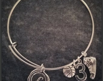 May include: Silver-toned bangle bracelet with multiple charms. Charms include a basketball, the number 3, a basketball shoe, and a circular charm with the letter "L". The bracelet is displayed against a dark gray background.