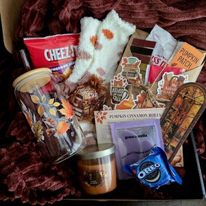 May include: A gift box containing a glass tumbler with a bamboo lid and autumn leaf print, fuzzy white socks with orange spots, a bag of Cheez-It crackers, a pumpkin spice candle, a pack of Oreo cookies, a bookmark, and various fall-themed stickers.
