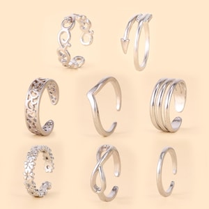 Könnte beinhalten: Eine Sammlung von acht silberfarbenen offenen Ringen mit verschiedenen Designs, darunter ein Blumenmuster, ein Unendlichkeitssymbol, eine Chevron-Form, ein dreireihiger Stil, ein Pfeilmotiv und komplizierte Ausschnittmuster auf hellem Grund.