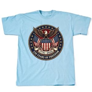 Może przedstawiać: Jasnoniebieski t-shirt z okrągłą grafiką przedstawiającą bielika amerykańskiego, dwie flagi USA i tarczę. Wzór zawiera wstęgę z napisem 1776-2026 oraz 250 YEARS OF FREEDOM, otoczoną obwódką ze złotych gwiazd.