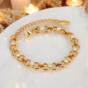 May include: A gold-toned chain bracelet featuring a series of interlocking circular links. Several of these links are accented with small, clear, round-cut stones embedded within the center. The bracelet includes a lobster claw clasp and an extender chain.