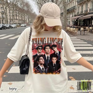 May include: An ivory short-sleeve t-shirt featuring a graphic print of five men in suits with the text ZHANG LINGHE in a bold arched font above them. The design includes a red and orange fiery background with small white star accents.