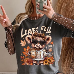 Op de afbeelding: Een antracietgrijs t-shirt met een afbeelding van een Schotse hooglander met een strik, zonnebril en een witte kabeltrui. De koe houdt een mok vast met de tekst Fueled by Chaos, omringd door herfstbladeren en zonnebloemen met de tekst Leaves Fall I Don't.