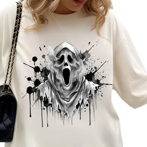 Könnte beinhalten: Ein cremefarbenes Langarmshirt mit einem schwarz-weißen Grafikdruck einer Kapuzenfigur mit schreiendem Gesichtsausdruck. Das Design enthält tintenartige Spritzer und Raucheffekte, die die zentrale Charakterillustration umgeben.