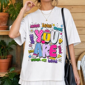 以下が含まれることがあります： 「YOU ARE」という大きな柄入りの文字がプリントされた、カラフルなグラフィックデザインの白い半袖Tシャツ。文字の周りには、鉛筆、リンゴ、電球、ハサミ、小さな星などの様々なイラストが描かれています。