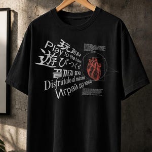 以下が含まれることがあります： 黒の半袖Tシャツ。円の中に赤い解剖学的な心臓のイラストが描かれ、Play to the future、玩到未来、遊びつくせ、끝까지 즐겨라、Disfrútalo al máximo、Играй до концаを含む多言語のテキストがプリントされています。