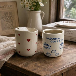 May include: Two off-white ceramic cups. The left cup features a notched rim and a pattern of small red hearts. The right cup displays the blue text "WANNA COFFEE?" alongside blue line drawings of a coffee cup, a mug, and three small cookies.
