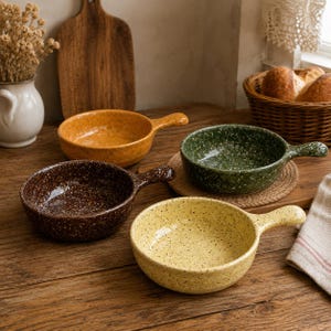 Puede incluir: Cuatro cuencos redondos de cerámica con asas, cada uno con un acabado moteado en colores distintos: amarillo mostaza, marrón oscuro, verde bosque y amarillo claro. Estos platos pequeños y poco profundos están diseñados para servir.