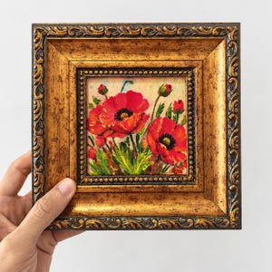 May include: A small square oil painting of vibrant red poppy flowers with green stems and leaves, set within an ornate, textured gold-colored frame. The artwork features detailed brushstrokes and a light-colored background behind the floral subject.
