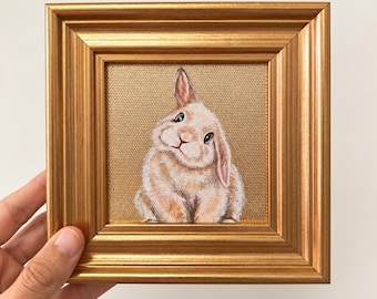 Puede incluir: Una pequeña pintura cuadrada de un conejo marrón claro con orejas largas y ojos oscuros, sobre un fondo dorado texturizado. La obra está encerrada en un marco decorativo de metal dorado con un diseño de borde estriado y escalonado.