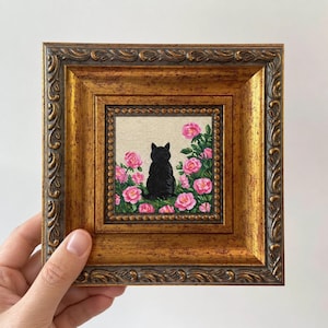 以下が含まれることがあります： 装飾的な金色のフレームに入った小さな正方形の絵画。背中を向けた黒猫が描かれており、明るいベージュのキャンバス背景に鮮やかなピンクのバラと緑の葉が添えられ、装飾的な金色の縁取りで仕上げられています。